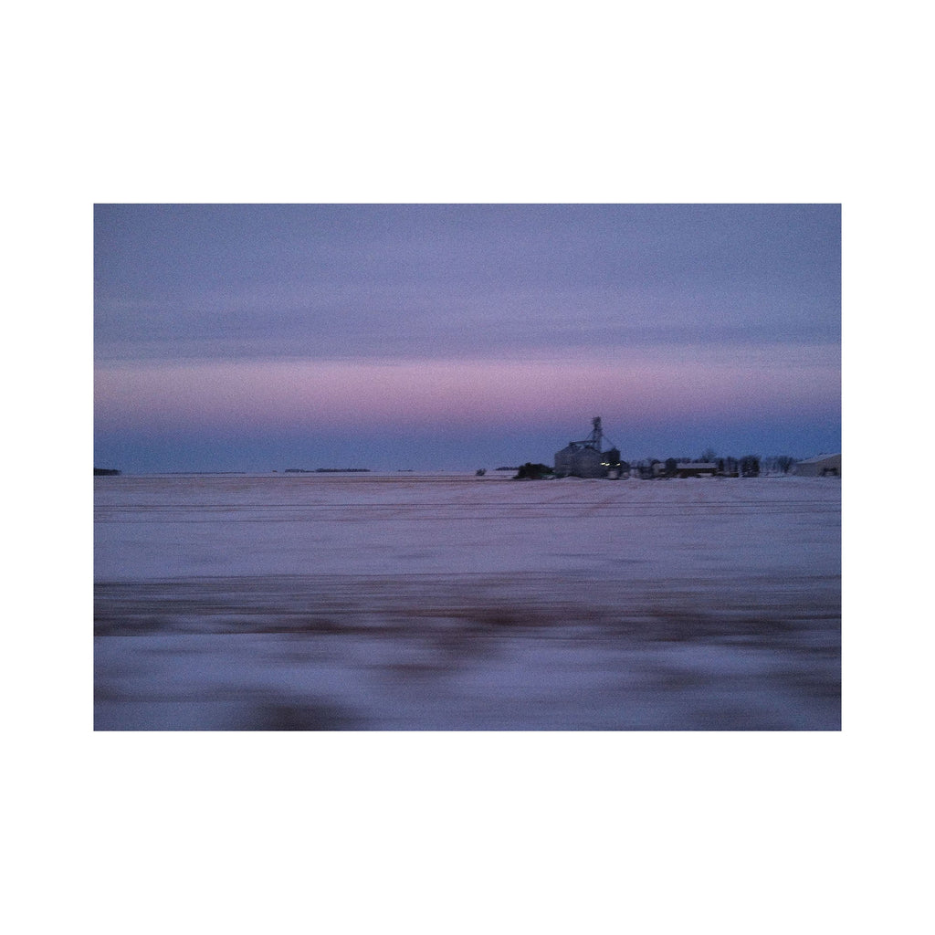 The artwork "Good Morning Indianna" by Kelly McGovern features a serene winter landscape with a vast snow-covered field under a soft, pastel-colored sky transitioning from light purple to blue. A distant farmhouse and silo are silhouetted against the horizon, creating a calm and quiet rural scene. The image evokes peaceful early morning stillness.