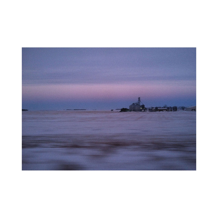 The artwork "Good Morning Indianna" by Kelly McGovern features a serene winter landscape with a vast snow-covered field under a soft, pastel-colored sky transitioning from light purple to blue. A distant farmhouse and silo are silhouetted against the horizon, creating a calm and quiet rural scene. The image evokes peaceful early morning stillness.
