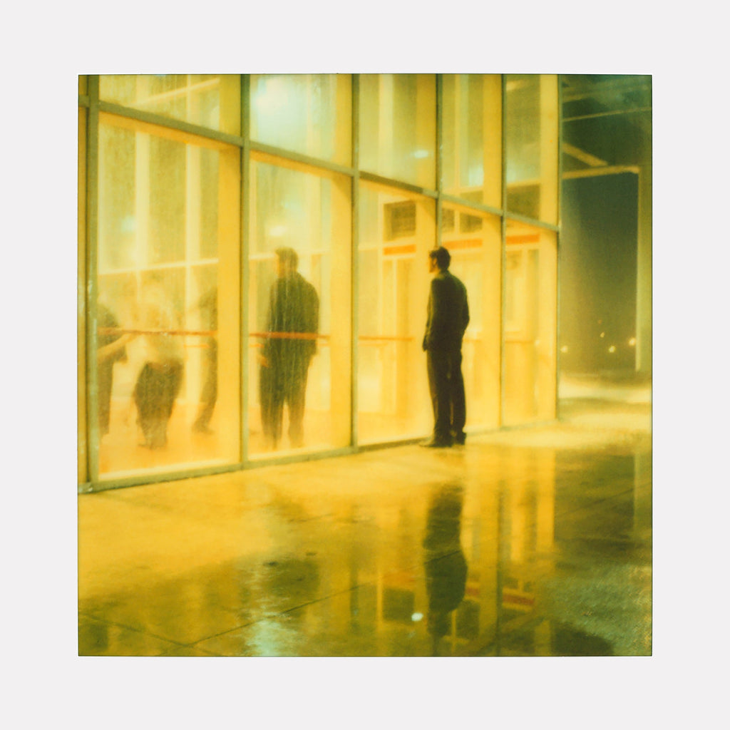 The artwork "Henry watching Athena Dance (Stay) - with Ryan Gosling" by Stefanie Schneider depicts a softly lit, reflective scene where a solitary figure stands outside a large glass window, observing blurred, dancing figures inside. Warm yellow tones and wet pavement reflections create a dreamy, atmospheric effect.