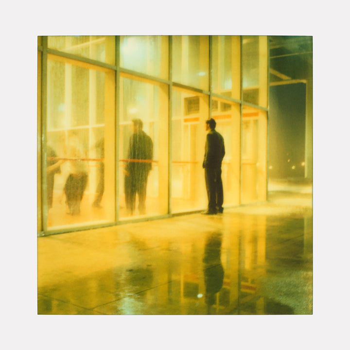 The artwork "Henry watching Athena Dance (Stay) - with Ryan Gosling" by Stefanie Schneider depicts a softly lit, reflective scene where a solitary figure stands outside a large glass window, observing blurred, dancing figures inside. Warm yellow tones and wet pavement reflections create a dreamy, atmospheric effect.