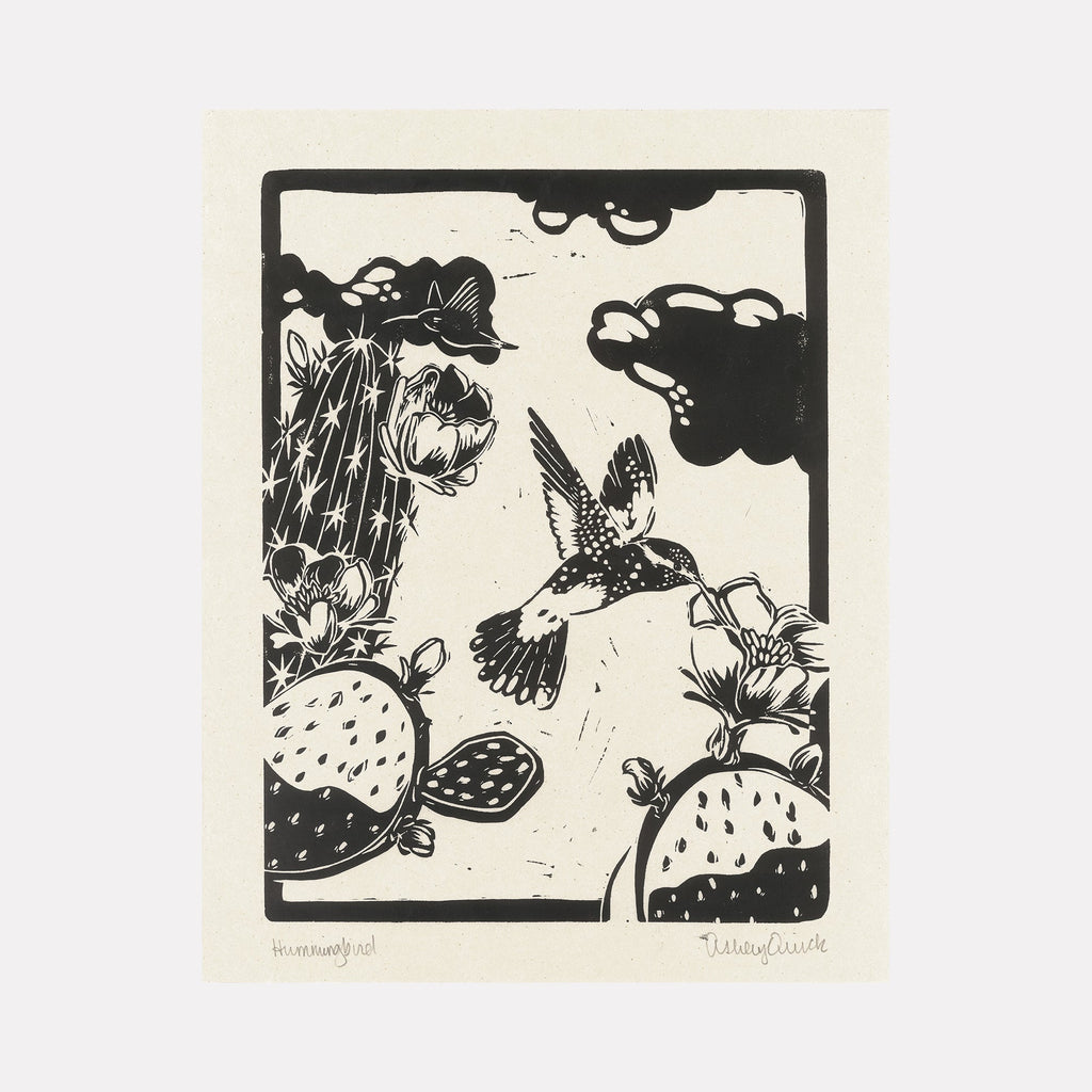 The artwork "Hummingbird" features a black and white linocut style print depicting a detailed hummingbird in mid-flight, surrounded by blooming cacti and stylized clouds within a rectangular frame. The composition emphasizes bold textures and organic shapes with a striking contrast. Ashley Ranae Quick.