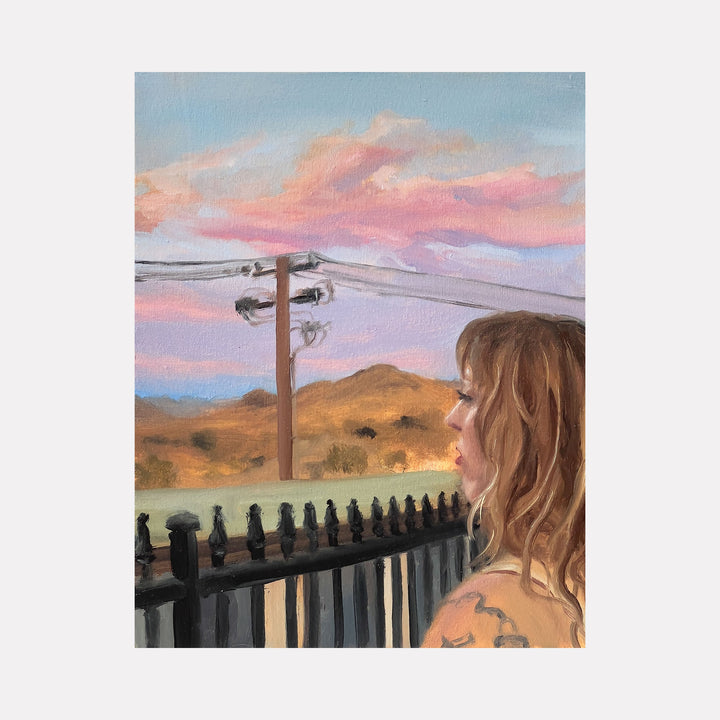 The artwork "Jen watching a Joshua Tree sunset" by Jillian Shea depicts a contemplative female figure gazing toward a pastel-hued sky with soft pink and lavender clouds above warm golden hills. The serene scene features detailed textures in hair and railing, evoking the intimate, light-infused style reminiscent of Edward Hopper and Gregory Thielker’s atmospheric realism.