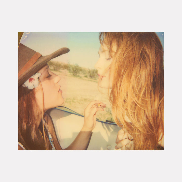 The artwork "Little Darlings (Till Death do us Part)" by Stefanie Schneider depicts two young girls with flowing hair, one wearing a wide-brimmed hat adorned with a flower, facing each other in soft, warm sunlight with a blurred natural background, evoking a nostalgic, dreamy atmosphere. Stefanie Schneider.