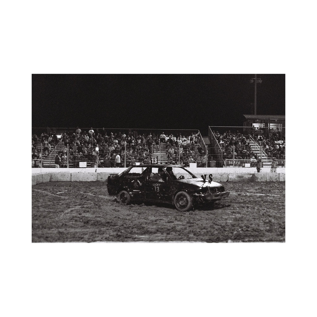 The artwork "Lucky 13" depicts a black demolition derby car with the number 13 painted on the door and roof, parked on a muddy arena track at night with a crowd of spectators seated on bleachers in the background. The monochrome image captures gritty textures and a rough, dynamic atmosphere. Kelly McGovern.