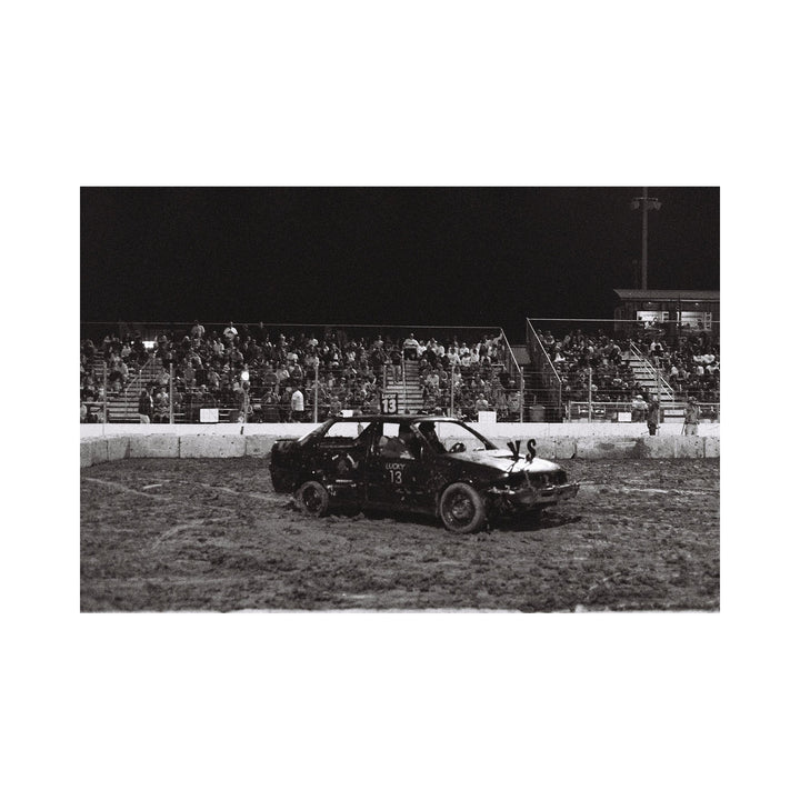 The artwork "Lucky 13" depicts a black demolition derby car with the number 13 painted on the door and roof, parked on a muddy arena track at night with a crowd of spectators seated on bleachers in the background. The monochrome image captures gritty textures and a rough, dynamic atmosphere. Kelly McGovern.