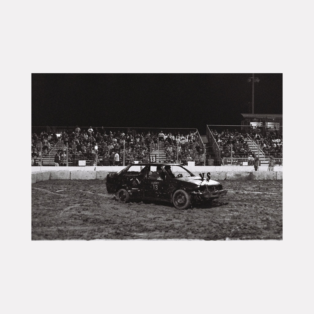 The artwork "Lucky 13" by Kelly McGovern captures a gritty black and white scene reminiscent of classic documentary photography with raw textures of dirt and metal. The image features a battered car marked with the number 13 at a racetrack, evoking the stark realism of artists like Dorothea Lange and modern street photographers, highlighting themes of endurance and grit.