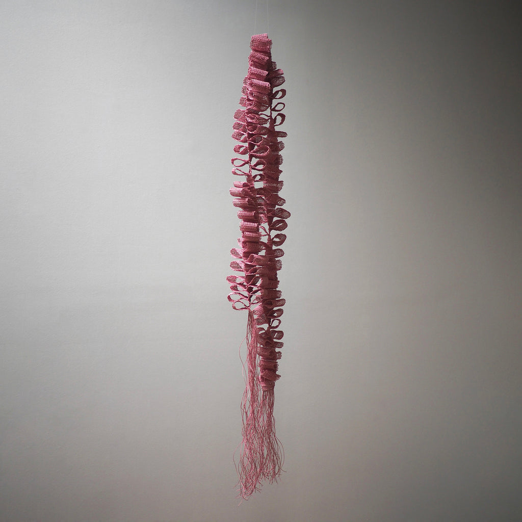 The artwork "Desire Paths" by Morgan Hale features a vertical, elongated sculpture composed of tightly coiled and looped pink fibers, resembling a delicate organic form. The intricate textures and flowing, fringe-like strands evoke the tactile qualities found in contemporary fiber art, reminiscent of works by Sheila Hicks and Eva Hesse.