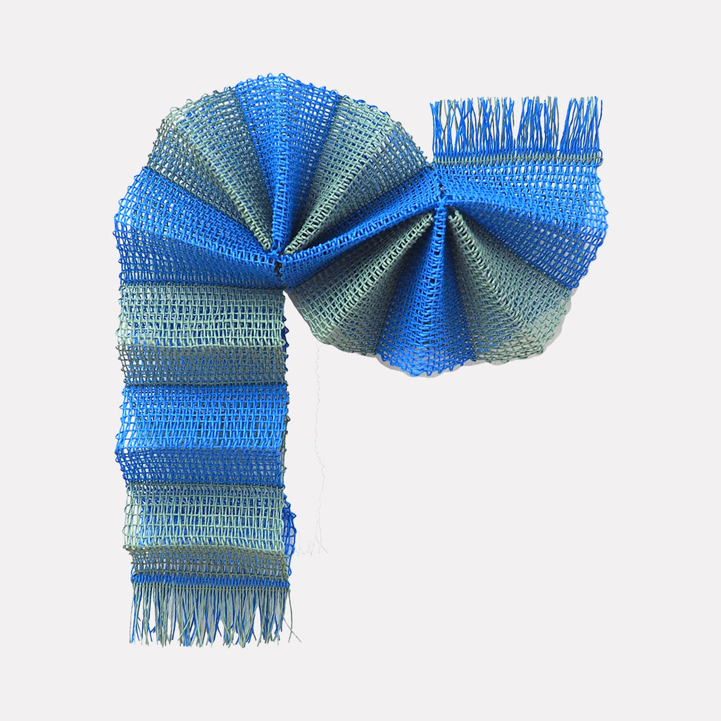 The artwork "Tree Tunnel" by Morgan Hale features a folded textile sculpture in vibrant blue and muted gray tones, showcasing a textured, woven pattern with layered loops and fringed edges. Its structured, twisting form evokes the dynamic compositions of artists like Anni Albers and the tactile minimalism seen in Sheila Hicks' fiber art.