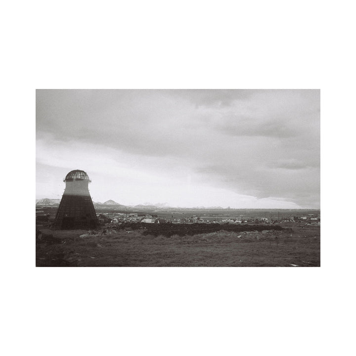 The artwork "Nowhere" by Kelly McGovern features a desolate landscape with a solitary, conical tower structure on the left side, set against a vast, cloud-filled sky. The scene is rendered in muted grayscale tones, emphasizing textures of rough earth and distant mountains, conveying isolation and stark natural beauty.