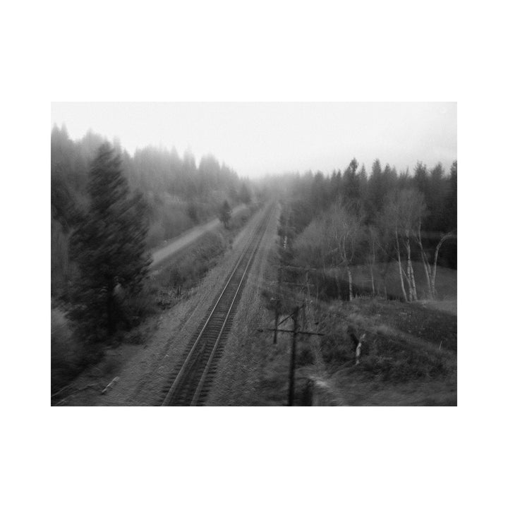 The artwork "One Day" by Kelly McGovern depicts a blurred black and white image of a railway track stretching into a foggy, forested landscape. The scene features bare and evergreen trees along a winding path beside the tracks, with a misty, atmospheric texture that evokes a sense of solitude and movement.