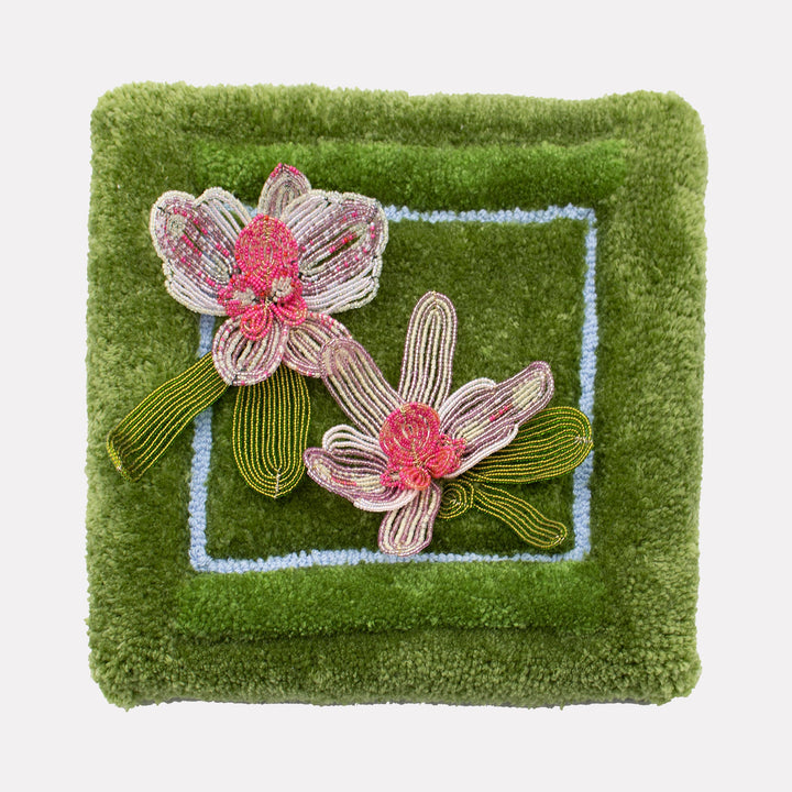 The artwork "Orchids" by Abigail Henthorne features two intricately beaded orchid flowers in shades of pink and white with detailed green leaves, set against a richly textured green and blue tufted background. The mixed media piece combines botanical delicacy with tactile depth, reminiscent of the floral studies by Georgia O’Keeffe and the decorative elegance of Klimt’s nature motifs.