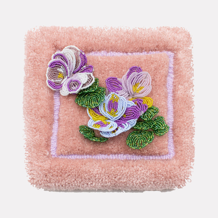 The artwork "Pansies" by Abigail Henthorne features a delicate mixed media composition with intricately beaded pansy flowers in shades of purple, white, yellow, and green, set against a textured, plush pink background. This tactile piece evokes the detailed craftsmanship reminiscent of textile art and the intricate natural forms found in Georgia O’Keeffe’s floral studies.