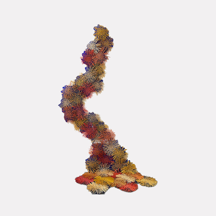 The artwork "RUSH" by Nora Chavooshian features a dynamic, vertical arrangement of textured, spiky floral shapes in warm tones of red, yellow, orange, and purple against a light background, creating a sense of organic movement and depth. The piece emphasizes intricate detail and vibrant color contrast. Nora Chavooshian.