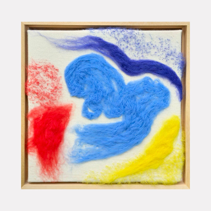 The artwork "RYB" features vibrant wool textures in primary colors with swirling and wispy forms of blue, red, yellow, and purple on a neutral background, creating a dynamic abstract composition. The piece is framed in light wood, emphasizing its tactile quality. By Ana Maria Farina.