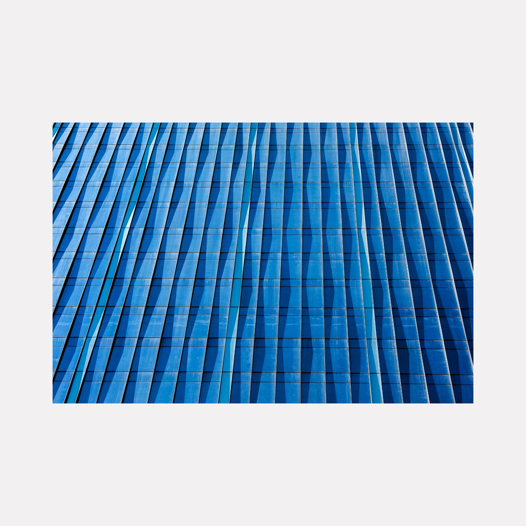 Reflections by Steve Edson captures the interplay of light and shadow on an architectural glass facade, featuring a rhythmic pattern of vertical lines in varying shades of blue that convey a sense of depth and dynamic movement through a modern, minimalist aesthetic.