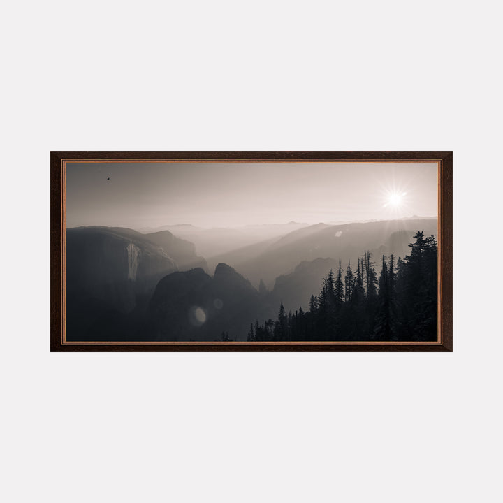 Remanoir by Kyle Murphy presents a tranquil black and white landscape of misty mountains fading into the horizon, with soft sunlight casting gentle highlights over tall pine trees in the foreground, evoking a serene and contemplative atmosphere that invites reflection on nature’s vastness and quiet beauty.