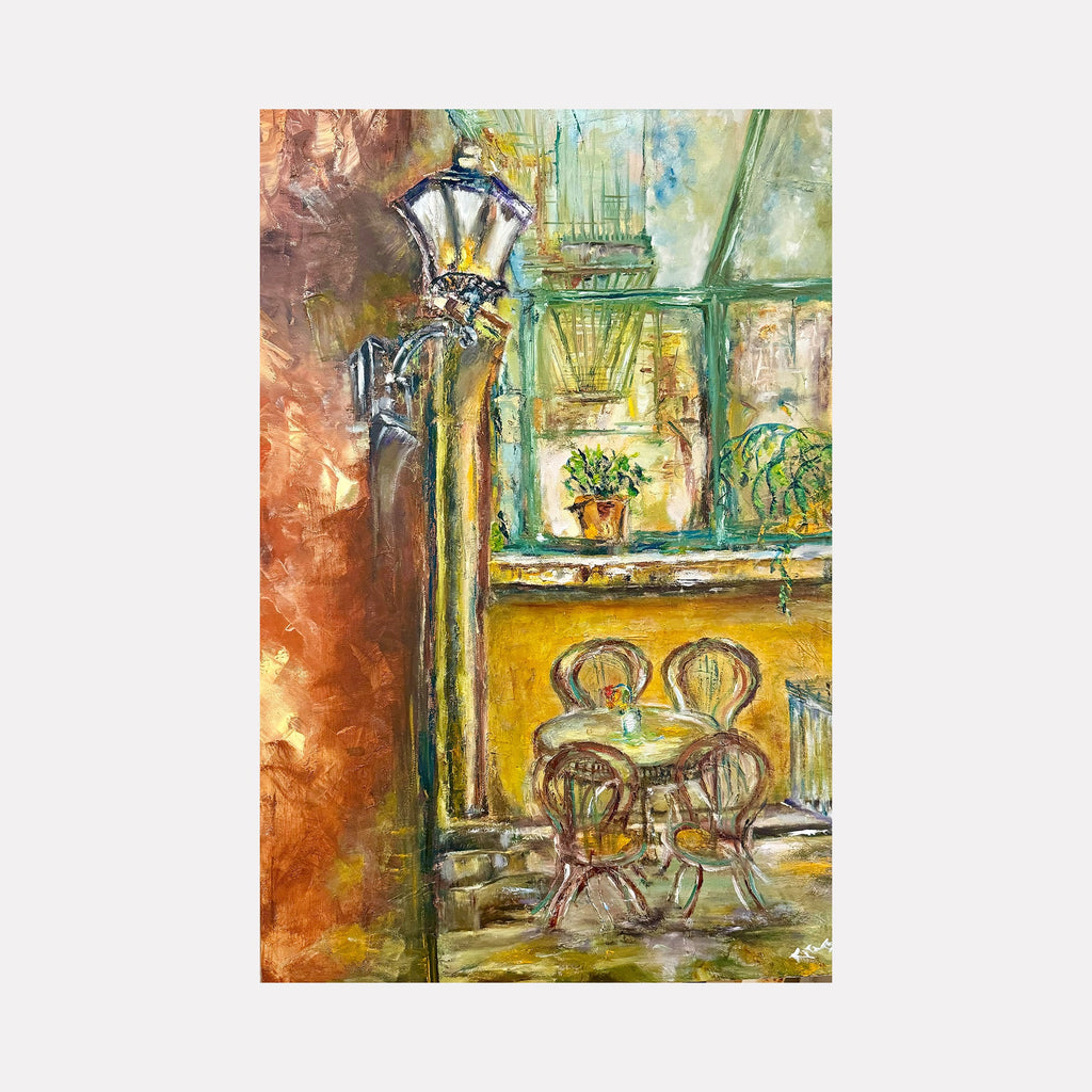 The artwork "La Lanterna-New York City" by Rita BasuMallick depicts a cozy outdoor café scene with a vintage street lamp illuminating a small table and three chairs. The painting showcases warm earthy tones with textured brushstrokes, blending golden yellows, greens, and browns to capture an inviting New York ambiance. Rita BasuMallick