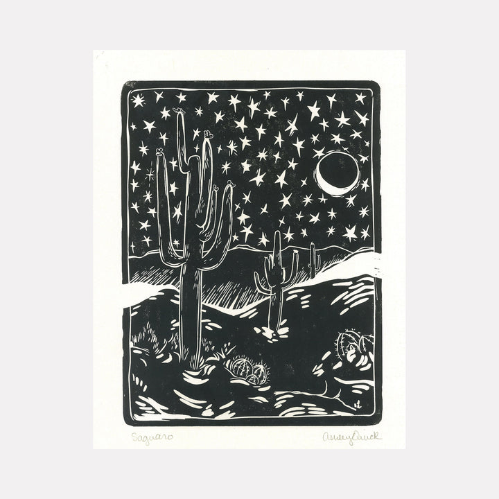 The artwork "Saguaro" features a stylized black and white desert landscape with tall saguaro cacti under a star-filled night sky and a crescent moon, rendered in a bold, graphic linocut style with contrasting textures and clean lines, by Ashley Ranae Quick.
