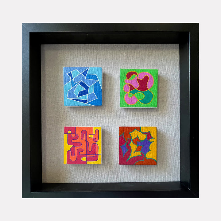 The artwork "Seasonal Phenomena II" by Joe Piscopia features four small square acrylic paintings on canvas with abstract geometric and organic shapes. Each piece showcases vibrant colors including blues, greens, pinks, yellows, reds, and purples, framed together in a sleek black frame. Joe Piscopia