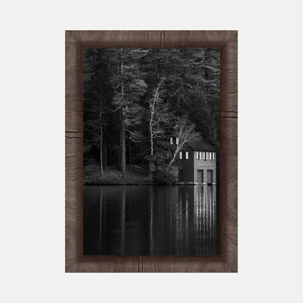 Shadows Rest by Kyle Murphy depicts a serene black and white scene of a quiet lakeside boathouse surrounded by dense, towering trees. The calm water perfectly reflects the structure and bare branches, creating a tranquil, contemplative mood within a natural woodland setting.