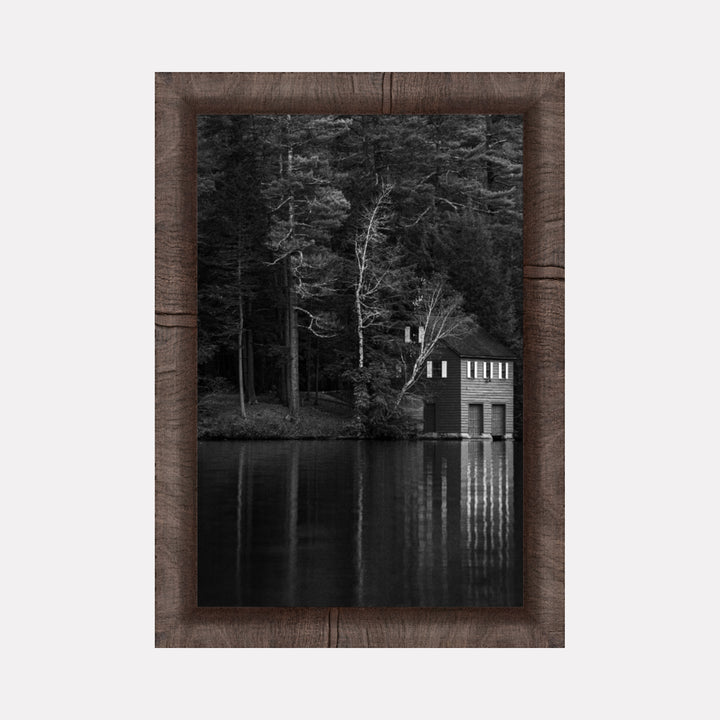 Shadows Rest by Kyle Murphy depicts a serene black and white scene of a quiet lakeside boathouse surrounded by dense, towering trees. The calm water perfectly reflects the structure and bare branches, creating a tranquil, contemplative mood within a natural woodland setting.