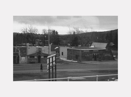 The artwork "Silient City" by Kelly McGovern depicts a quiet, desolate urban scene in grayscale, emphasizing stark contrasts and muted textures reminiscent of Edward Hopper’s realism and the somber mood of early 20th-century photography. The composition captures empty buildings, barren trees, and a solitary figure, evoking themes of isolation and stillness.