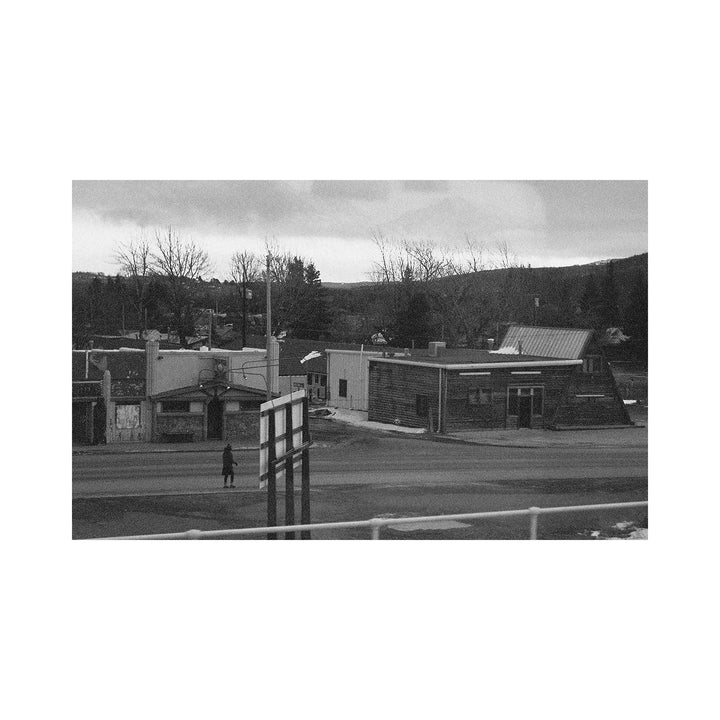 The artwork "Silient City" features a black and white image of a quiet, desolate urban scene with empty streets, simple industrial and residential buildings, bare trees, and cloudy skies, evoking a sense of stillness and solitude. The textured grayscale tones and stark contrasts emphasize the somber mood. By Kelly McGovern.