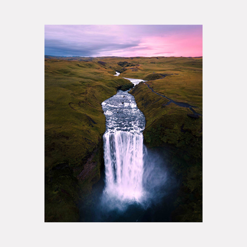 The artwork "Skógafoss" by Kyle Murphy in gallery position 1 presents a dramatic aerial view of a powerful waterfall cascading through a rugged, moss-covered landscape. The soft, pastel pink sky contrasts with the vibrant green terrain, evoking the immersive natural detail found in Albert Bierstadt’s luminous landscapes and the atmospheric mood reminiscent of Caspar David Friedrich’s romantic scenes.