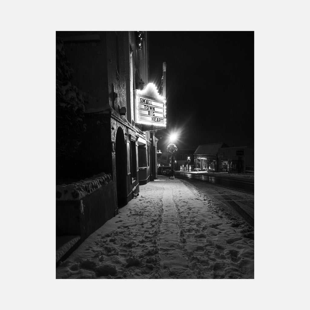 Small Town, Big Heart by Dennis Maida: Black and white photograph capturing a snowy evening streetscape featuring an illuminated storefront sign casting a warm glow onto snow-covered sidewalks. The atmospheric composition shows a vintage small-town setting with classic architecture and street lamps creating dramatic contrast in the night scene.