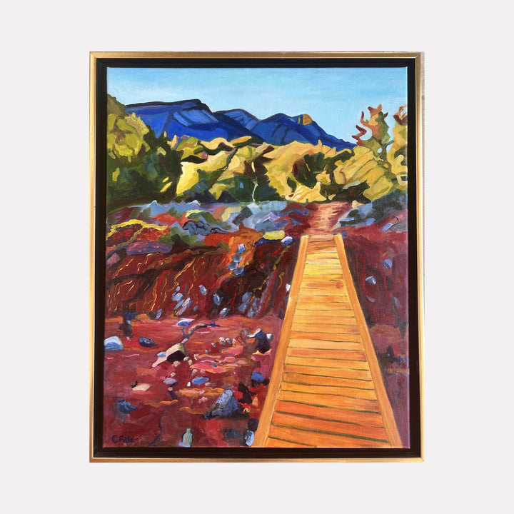 The artwork "Strip MIne Trail 6" by Connie Falk depicts a vibrant landscape featuring a wooden boardwalk leading through a rich, earthy terrain with red and brown tones, accented by scattered rocks. The background reveals lush green trees and distant blue mountains under a clear sky, rendered in a style reminiscent of early 20th-century American modernists like Georgia O’Keeffe and the bold colors of Fauvism.