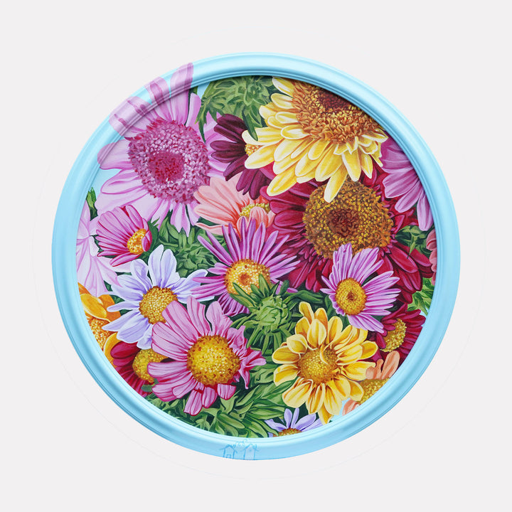 The artwork "Sun" by Carlos Gamez de Francisco displays a vivid array of daisies in shades of pink, yellow, and purple, surrounded by lush green foliage. The detailed floral composition and vibrant palette evoke a cheerful, natural warmth that celebrates the lively energy of a blooming garden.