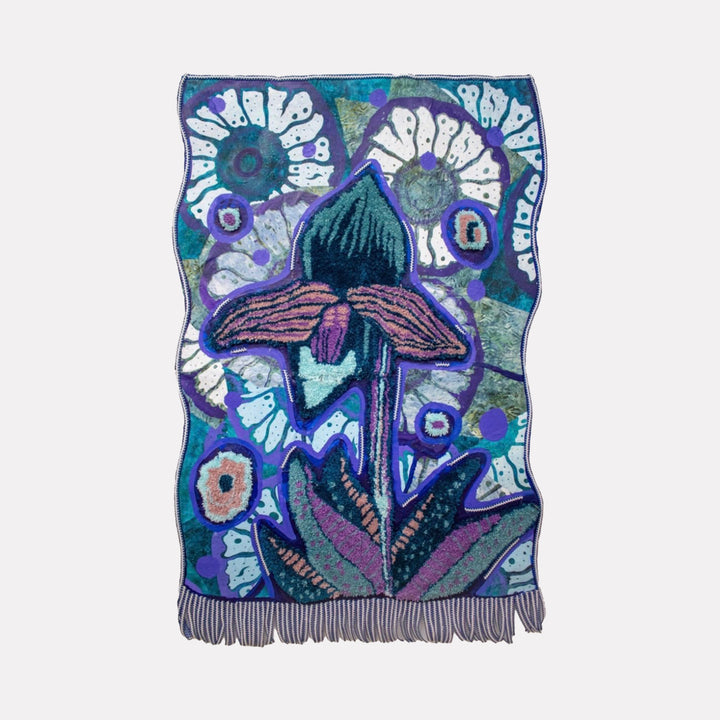 The artwork "Surprise in Blue" features a stylized floral design with a large central flower in shades of blue, purple, pink, and teal, set against a textured background with circular and abstract shapes in white and blue. The piece has a woven texture with fringed edges, showcasing an artistic mix of organic and geometric elements, by Laurie Shapiro.