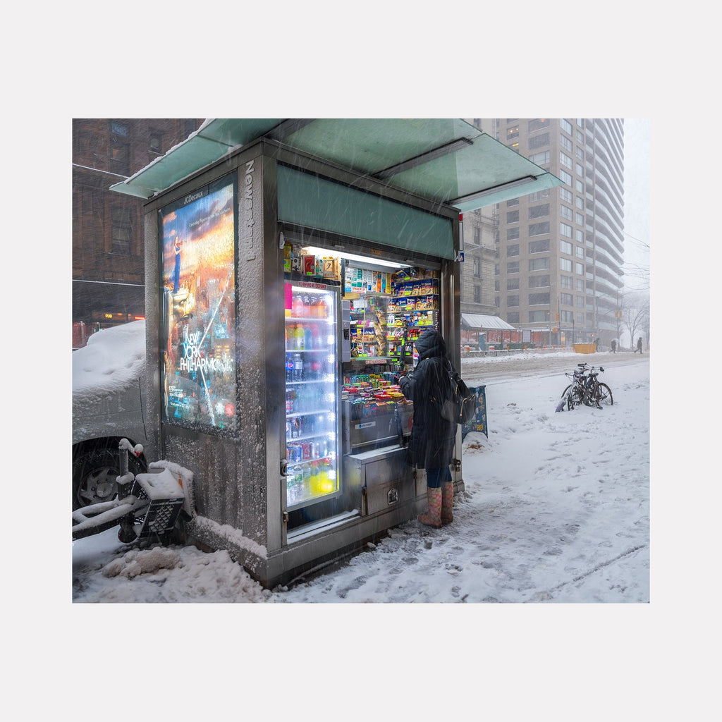 The artwork 'Urban Soles' by Dennis Maida captures a city newsstand illuminated at dusk during a snowstorm. A vending kiosk with vibrant neon lights and colorful product displays stands against winter-grey urban backdrop of high-rise buildings. Snow blankets the ground while a bicycle rests nearby. The photograph's composition contrasts warm artificial lighting with cool winter tones, creating an atmospheric urban scene.