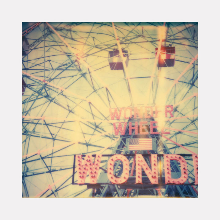 The artwork "Wonder Wheel (Stay)" showcases a vintage Ferris wheel structure with intricate metal beams and cabins, bathed in soft, warm light with a nostalgic, faded color palette. The illuminated sign with large letters reads "Wonder Wheel" above an American flag, capturing a retro amusement park atmosphere. By Stefanie Schneider.