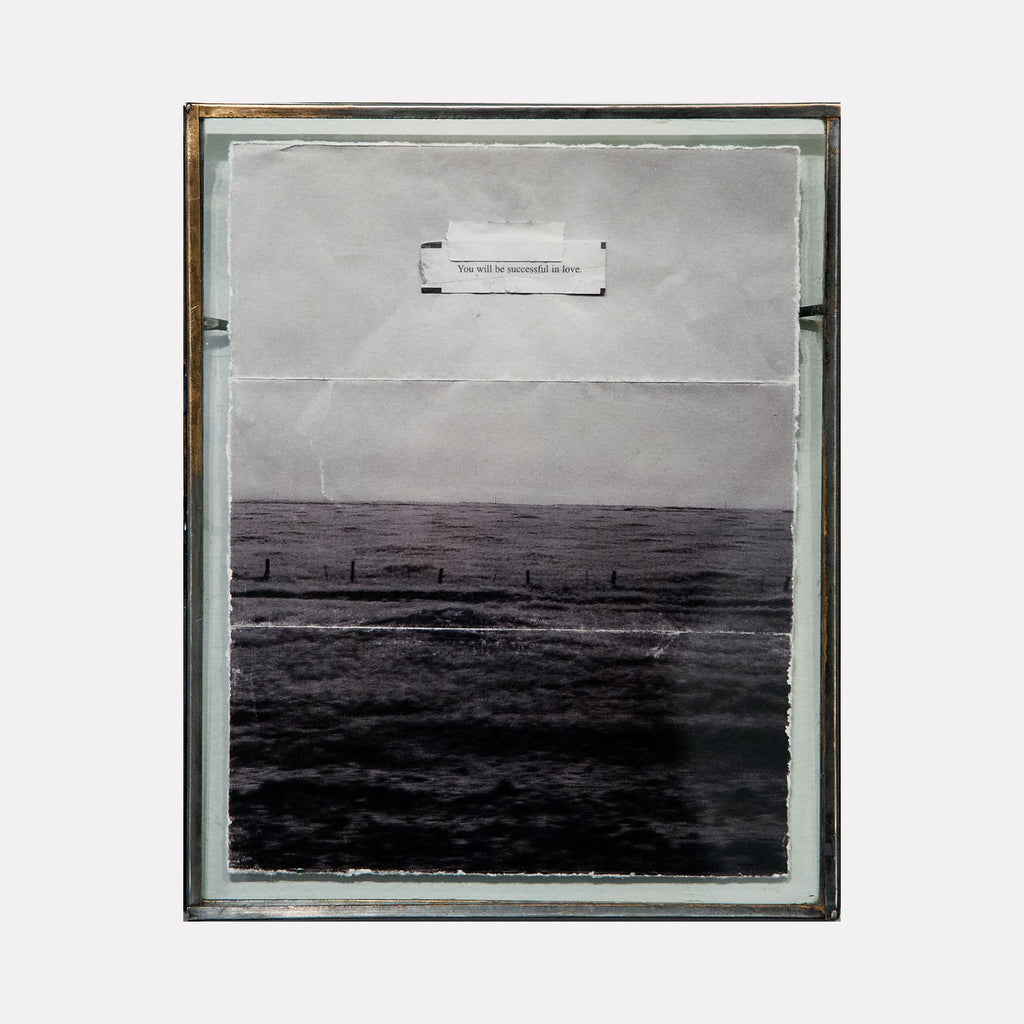 The artwork "You Will be Successful in Love" by Kelly McGovern features a layered collage style with a monochromatic palette of grays and blacks, evoking a serene yet somber ocean scene. The top section showcases a torn paper with typed text, reminiscent of conceptual works by Robert Rauschenberg, merging texture and message in a delicate, minimalist composition.
