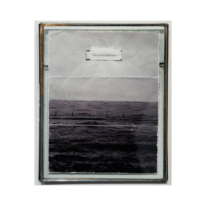 The artwork "You Will be Successful in Love" by Kelly McGovern features a textured, grayscale collage with an image of a dark, rippling sea on the lower half and a crumpled paper background above. A small, typed phrase "You will be successful in love" is centered near the top, framed by a simple, thin metallic border. Kelly McGovern.
