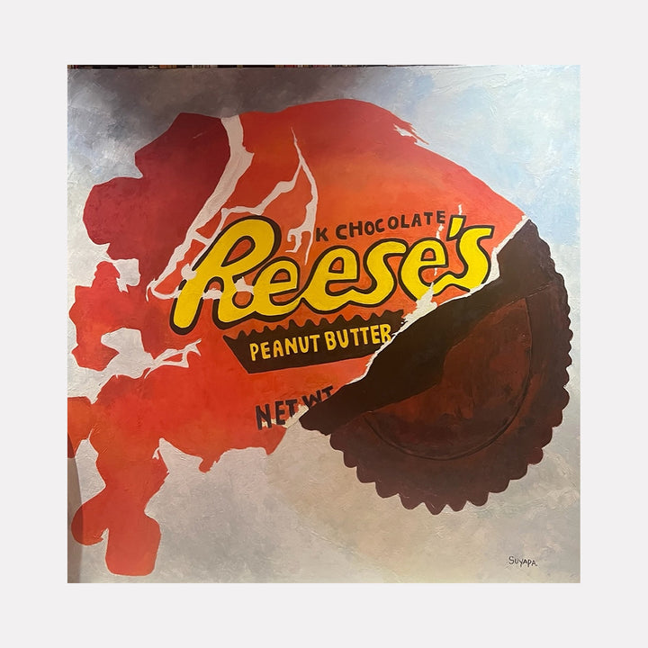 The artwork "Finally Here" by Suyapa Quinn features a large, partially unwrapped Reeseâ€™s Peanut Butter Cup against a textured background blending soft blues and grays. The bold orange and brown candy wrapper contrasts with the realistic, slightly abstracted form, evoking pop art influences reminiscent of Andy Warholâ€™s iconic commercial imagery and Claes Oldenburgâ€™s playful consumer culture sculptures.