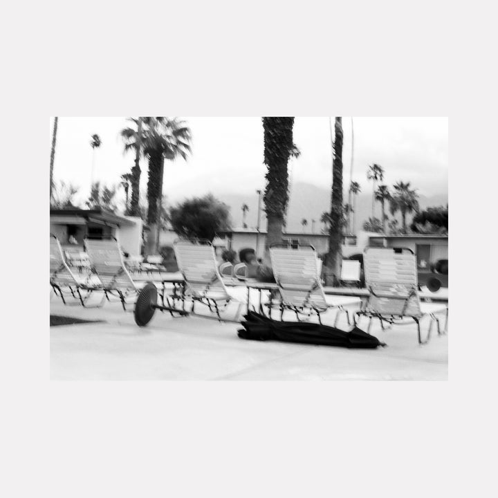 The artwork 'blackwind' captures empty pool loungers and palm trees in black and white film photography, creating serene desert ambiance by Esther Voisin