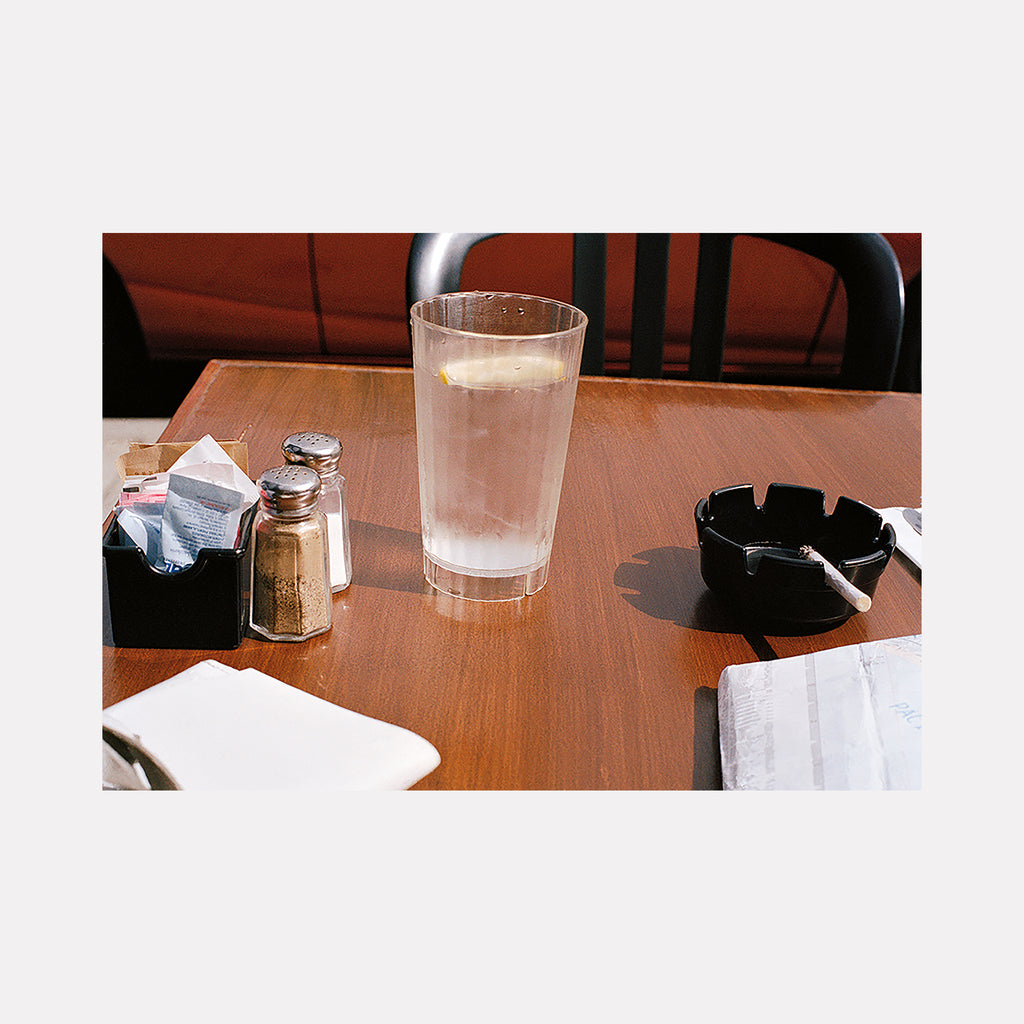 The artwork 'melrose' captures a diner table scene with glass of water, salt shaker, and black ashtray on wooden surface in film photography by Esther Voisin