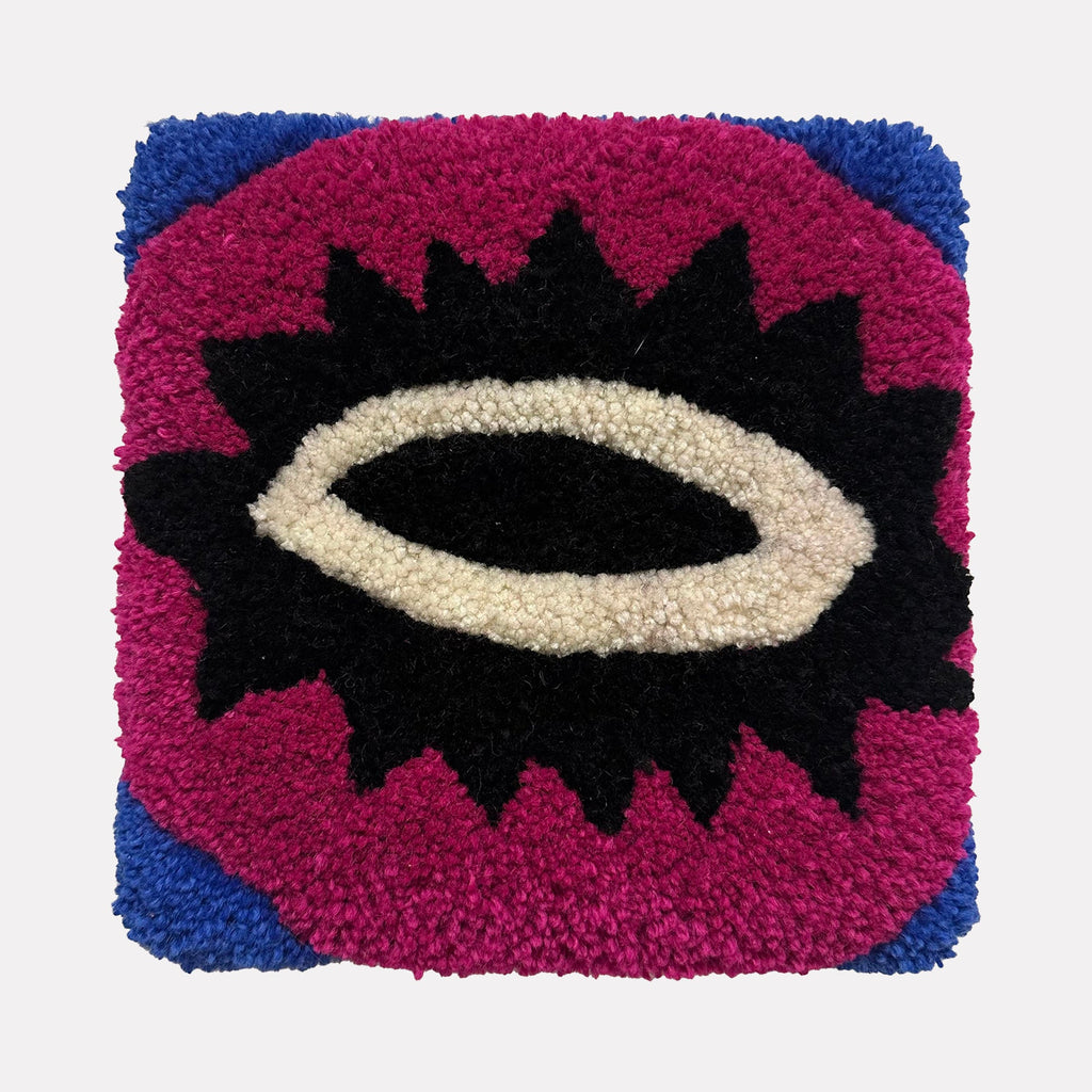 The artwork "Olho" by Ana Maria Farina features a textured, tufted square design with bold colors. It showcases a central eye-like shape in black and white, surrounded by a spiked black outline on a vibrant magenta background with deep blue corners. The piece highlights a tactile, handcrafted quality.