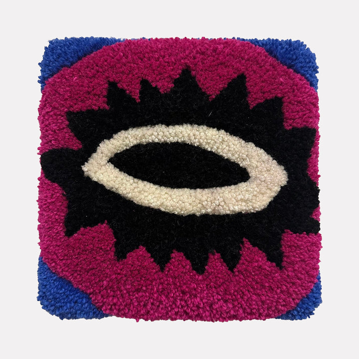 The artwork "Olho" by Ana Maria Farina features a textured, tufted square design with bold colors. It showcases a central eye-like shape in black and white, surrounded by a spiked black outline on a vibrant magenta background with deep blue corners. The piece highlights a tactile, handcrafted quality.