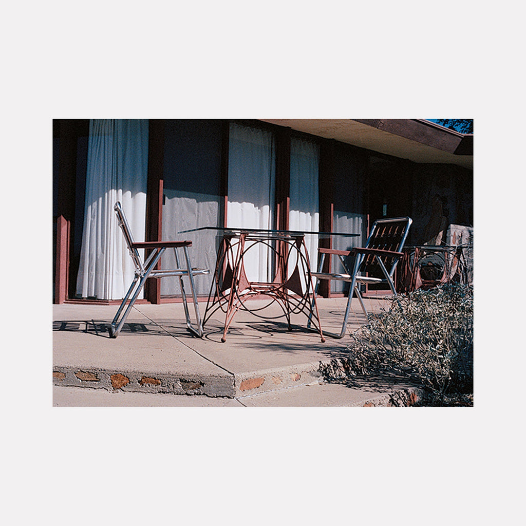 The artwork 'two' captures vintage metal chairs and table on sun-lit motel patio, white curtains behind, muted retro aesthetic by Esther Voisin