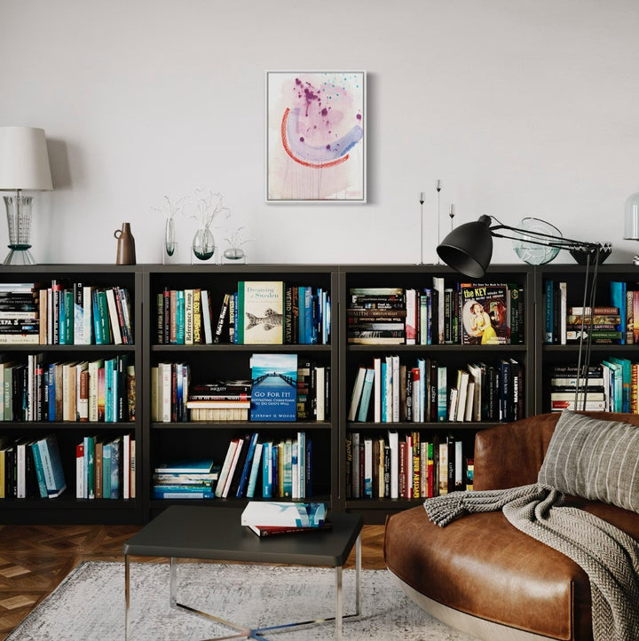 The artwork 'The Twins, No.1' by Taylor Binda showcased above a modern bookshelf features abstract splashes of purple and blue, accented with a sweeping coral arc. Created with acrylic, coffee and oil pastel on watercolor paper, the framed piece creates a striking focal point against the neutral wall, complementing the contemporary reading space.