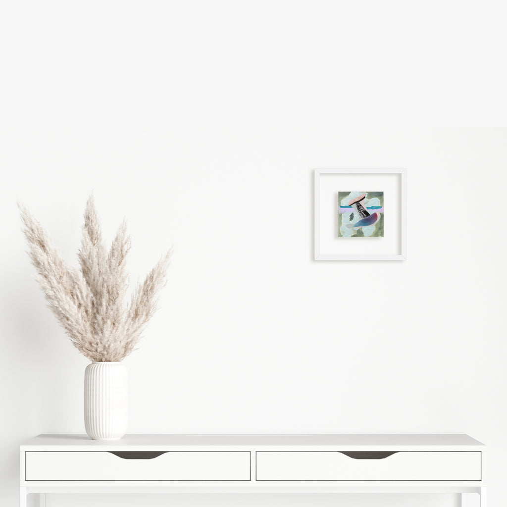 The artwork 'Fragment#01' by Winnie Sidharta features abstract shapes in soft blue and purple tones created with watercolor and magazine collage elements, displayed in a white frame. The 5x5 inch mixed media piece hangs above a minimalist white console table decorated with dried pampas grass in a ribbed ceramic vase.