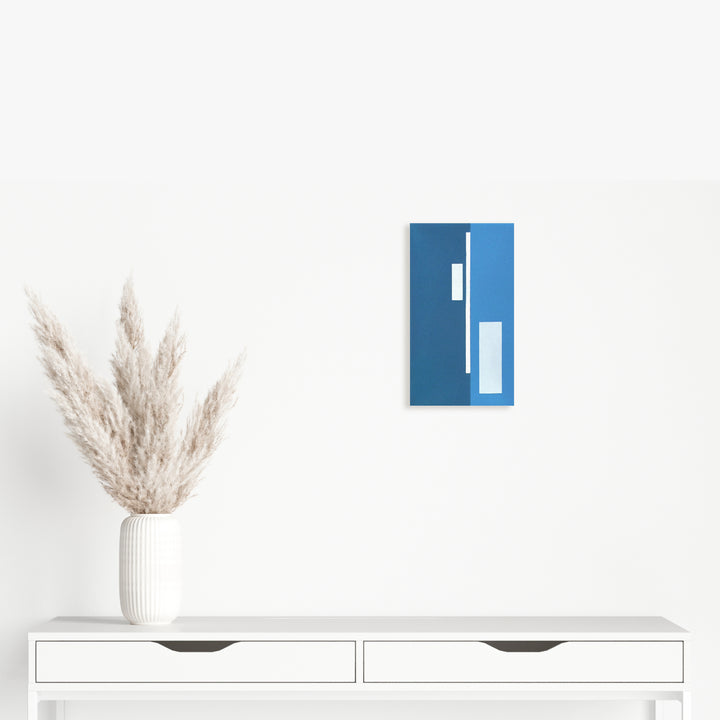 The artwork 'Moments II (Triptych)' by Shyun Song features a minimalist geometric composition on blue canvas displaying white rectangular shapes arranged vertically, accompanied by a decorative pampas grass arrangement in a white ribbed vase positioned on a modern white console table.