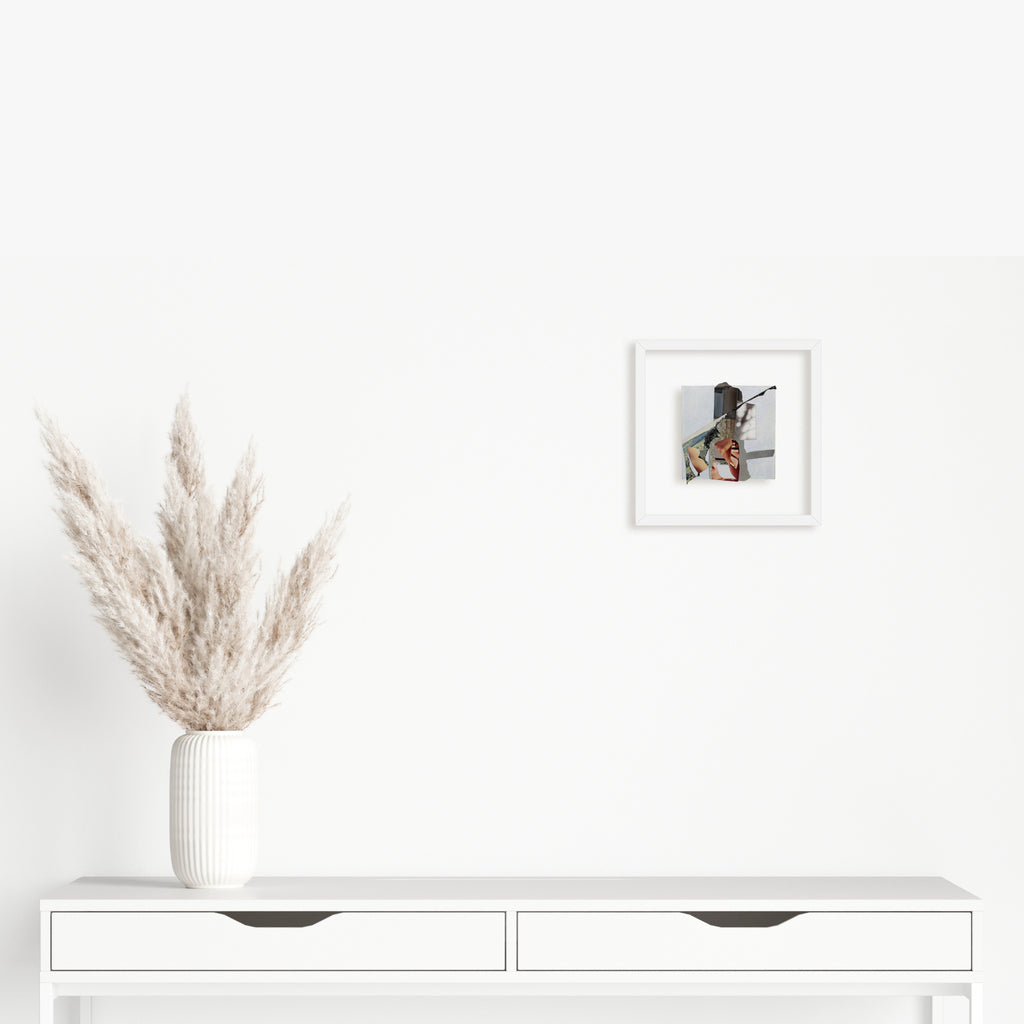 The artwork 'Fragment#02' by Winnie Sidharta features a delicate watercolor piece with vibrant magazine collage elements set within a pristine white frame. The 5x5 inch panel displays a modern mixed-media composition alongside decorative pampas grass in a white vase on a minimalist console table.