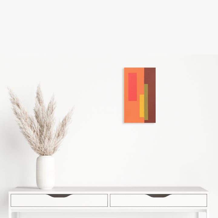 The artwork 'Moments III (Triptych)' by Shyun Song features bold geometric color blocks in warm tones of coral, terracotta, yellow, and burnt orange arranged vertically on canvas. The minimalist composition is displayed above a white modern cabinet with pampas grass in a ribbed white vase, creating a contemporary aesthetic.