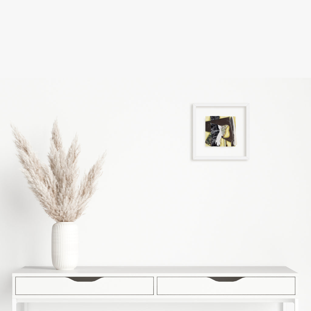 The artwork 'Fragment#06' by Winnie Sidharta features a striking mixed media composition displayed in a white frame, showcasing an abstract collage combining watercolor and magazine cutouts. Black and yellow tones create dynamic contrast on a 5x5 inch panel, presented above a minimalist white console with pampas grass decor.