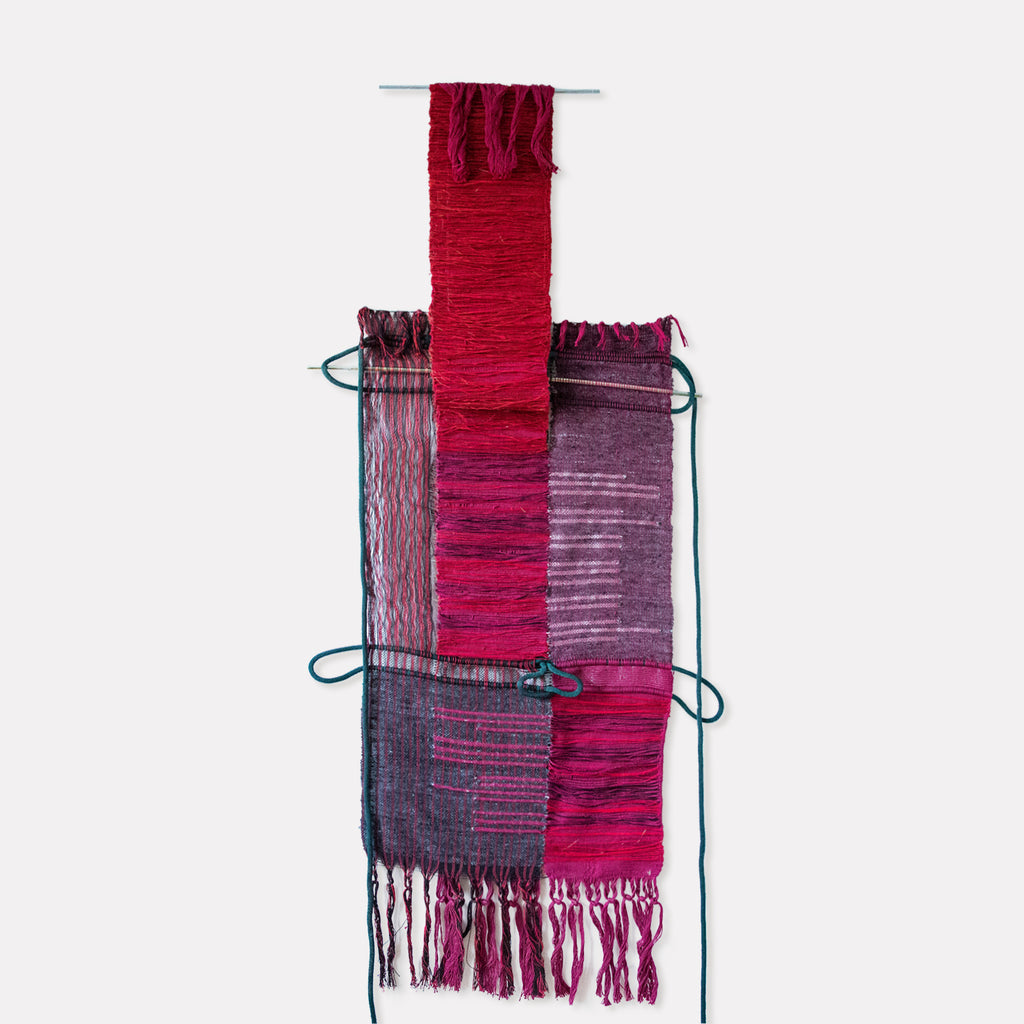 The artwork 'Slow Rising, English Morse Code' by Kathie Halfin features a vertical textile composition with vibrant crimson and purple hues. Hand-woven hemp and cotton create textural contrasts, with silver floss yarn details and copper wire accents. The piece displays a striking central red column intersecting horizontal purple bands, finished with delicate fringe tassels and teal binding elements.