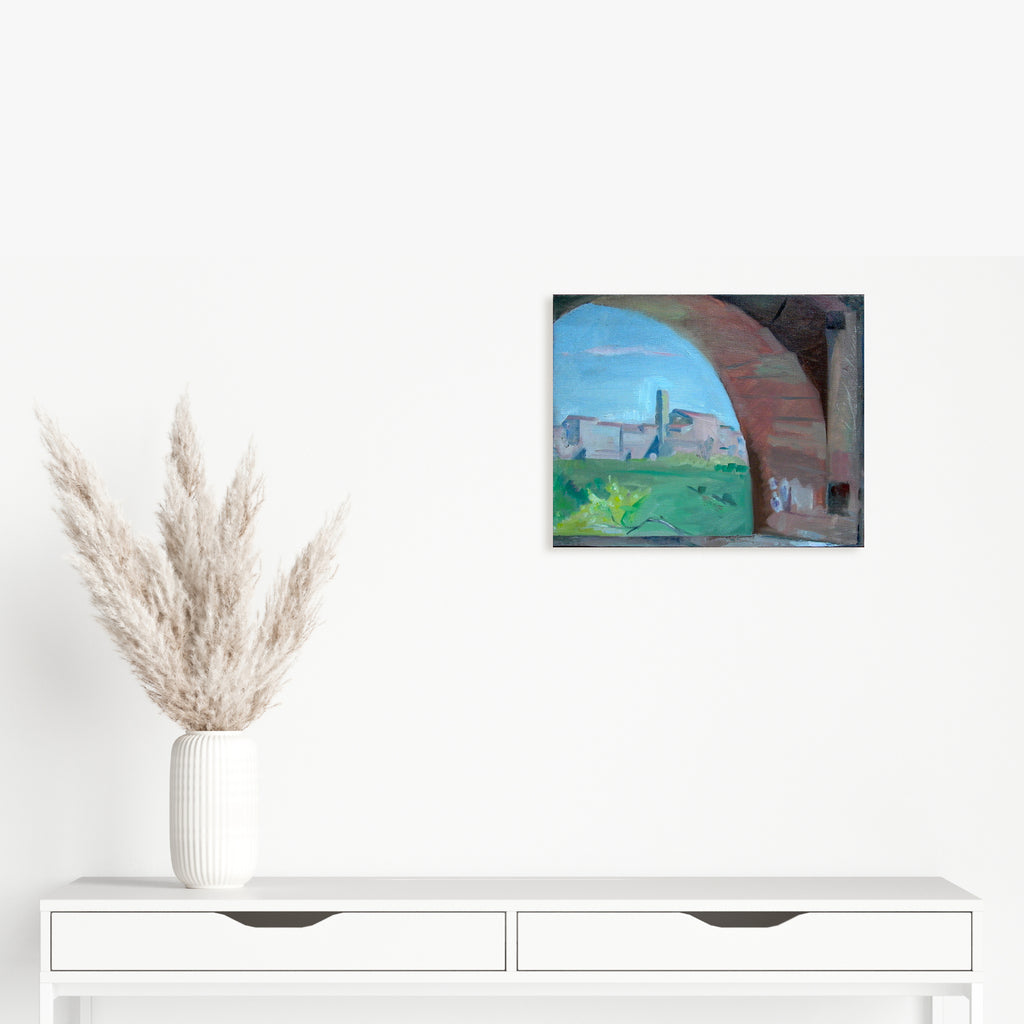 The artwork 'Through the Arch, Casole' by Peter Colquhoun captures a scenic Italian landscape viewed through a terracotta stone archway. The composition features vibrant blue skies, distant medieval buildings on a hilltop, and lush green countryside, all framed by the rustic arch's curved stonework. Oil on linen, 13x16 inches.